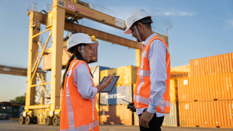 Construction technology trends contractors should be watching Two workers in hard hats and orange vests review notes on a tablet at a shipping container yard