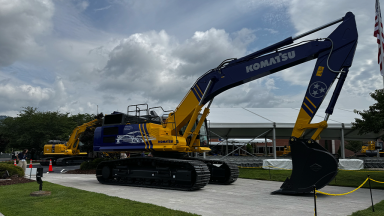 Large blue and yellow Komatsu excavator on display outdoors with another machine in the background near a white tent.