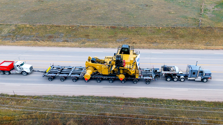 Aerial view of a large yellow construction machine being hauled on a multi-axle flatbed trailer pulled by trucks on a highway, with farmland nearby.