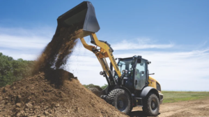 Backhoe loader dumping soil into a large dirt pile at a construction site under a blue sky.