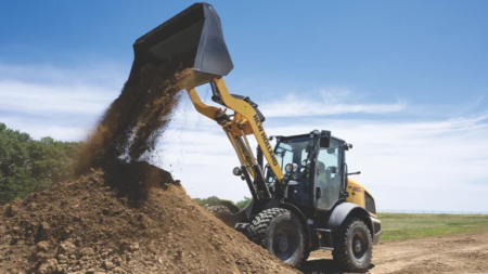 1784320718_64019_en_62d16_66186_new-holland-w80d-wheel-loaderpng - Machines Wiki Backhoe loader dumping soil into a large dirt pile at a construction site under a blue sky.