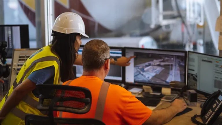 Two control-room staff monitor multiple screens, one pointing at the displays, in an industrial facility.”] ,