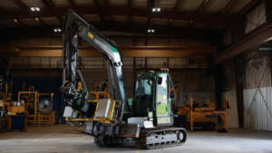 Compact excavator inside a large workshop lifting a metal crate with its hydraulic arm, with yellow machinery visible in the background,