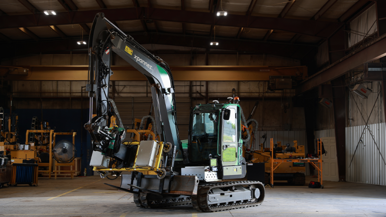 VIDEO: Xpanner launches excavator-based solar panel installer Compact excavator inside a large workshop lifting a metal crate with its hydraulic arm, with yellow machinery visible in the background,
