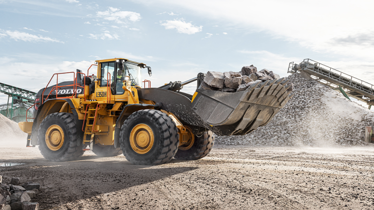Volvo CE’s Q2 report reflects strong organic growth amid market expansion Yellow Volvo wheel loader scoops rocky material at a quarry, moving a large bucket of crushed stone toward a rock pile under a blue sky