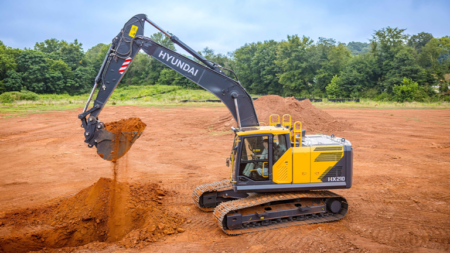 1784733533_64040_en_57639_66220_hyundai-hx210l-excavator-1png - Machines Wiki Yellow Hyundai excavator on a dirt construction site digging a trench with a pile of soil nearby and trees in the background, mid‑dig action.