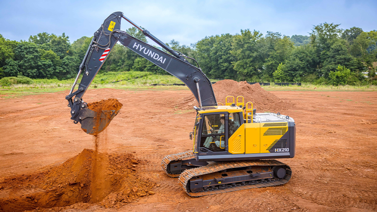Yellow Hyundai excavator on a dirt construction site digging a trench with a pile of soil nearby and trees in the background, mid‑dig action.