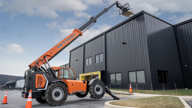 1784833246_64043_en_a45ce_66223_jlg-skytrack-telehandlerpng - Machines Wiki Orange SkyTrak telescopic forklift with an elevated work platform near a dark metal warehouse, safety cones in the foreground.