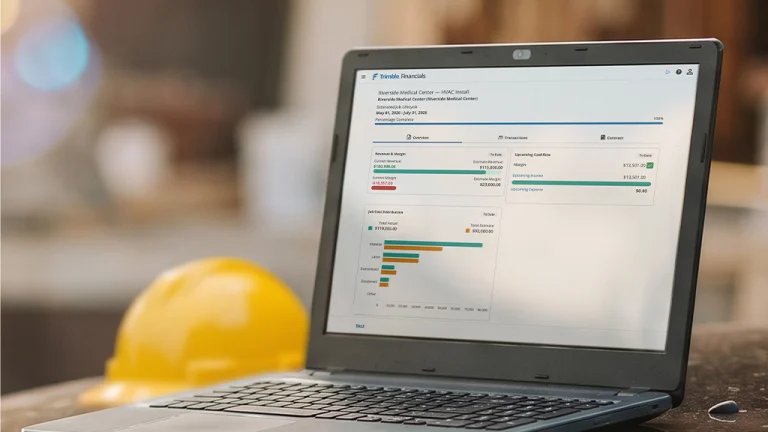 Laptop screen shows a financial dashboard with charts and figures, yellow hard hat blurred in the background on a desk.