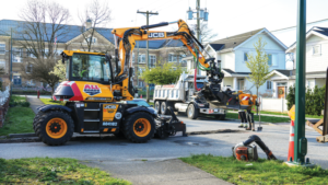 Orange JCB excavator lifting material into a dump truck on a residential street as workers supervise, with a curb, sidewalk, and houses in the background and a hose reel on the ground nearby.