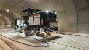 Wirtgen tunnel boring machine operating inside a curved concrete tunnel, with orange crawler tracks and hoses connected to the unit.