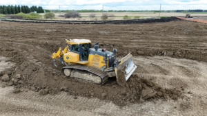 Yellow tracked excavator with a front blade moving soil on a large construction site; dirt mounds and cleared fields in the background.