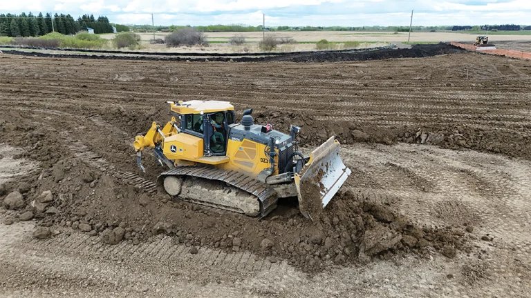 Yellow tracked excavator with a front blade moving soil on a large construction site; dirt mounds and cleared fields in the background.