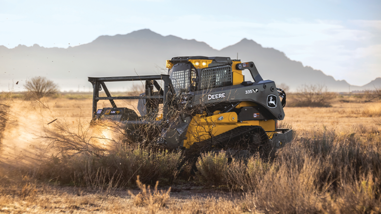 John Deere tracked skid-steer loader with a brush attachment clearing dry brush in a desert field, mountains in the distance.