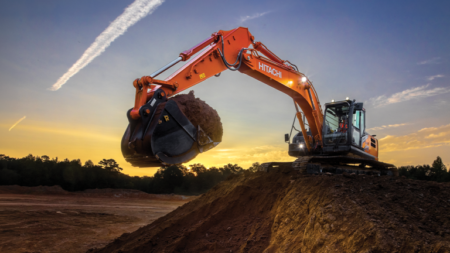 Orange Hitachi excavator on a dirt hill at sunset, lifting a large bucket of soil