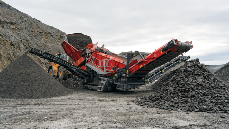 Industrial rock crusher and conveyor system at a quarry, processing stone into gravel with piles of crushed rock nearby