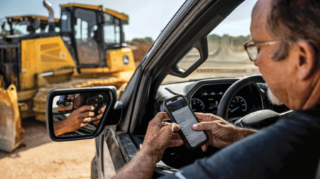 Man in a pickup at a construction site checks his smartphone; a yellow excavator sits in the background.