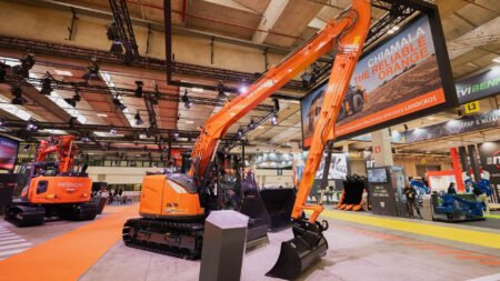 Large orange excavator on display at an indoor construction equipment trade show, with hanging rigging and a banner advertising a model or brand.