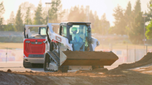 1785446731_64105_en_419e1_66295_bobcat-ssl-x-gamespng - Machines Wiki A compact skid-steer loader with a dirt bucket moves soil at a construction site, with a larger vehicle visible in the background.