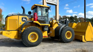 1785461697_64096_en_9e483_66287_afex-john-deere-wheel-loaderpng - Machines Wiki Yellow John Deere wheel loader with a front bucket and snow plow attachment parked on a paved lot, under a blue sky.