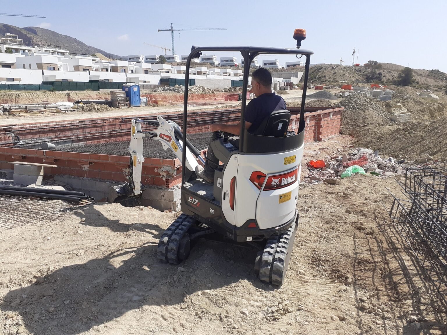 Operator sits in a Bobcat compact excavator on a dirt construction site with rebar and piles of rubble nearby.