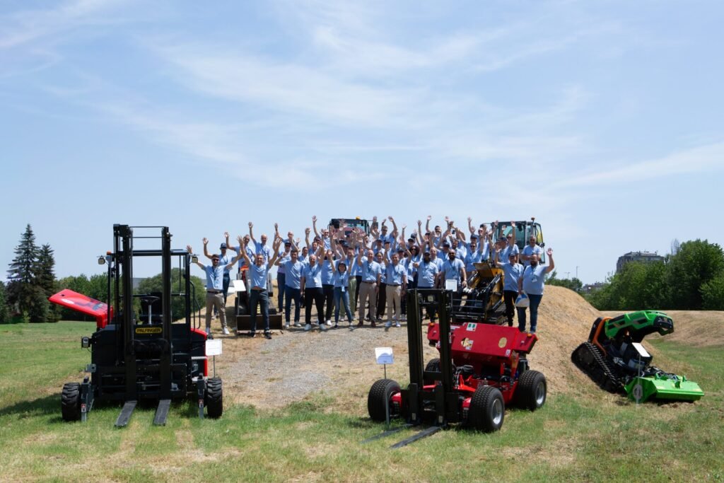 Group of people in blue shirts waving from behind large farm equipment outdoors on a sunny day.