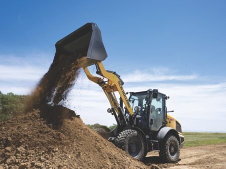 727967jpg - Machines Wiki Excavator dumping soil from its bucket onto a large dirt pile at a sunny construction site.