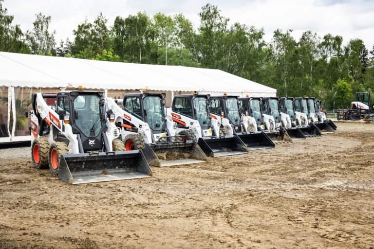 Row of Bobcat skid-steer loaders with buckets, lined up in front of a large white tent on a dirt lot outdoors.