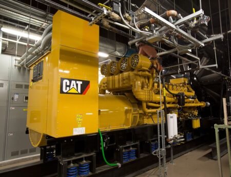 Large yellow CAT diesel generator set with exhaust piping and control panel in an industrial room.