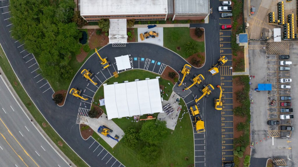 Komatsu’s Chattanooga Facility Celebrates 40 Years and Unveils New Machines Aerial view of a construction yard with several yellow cranes surrounding a white tent on a curved asphalt driveway near a building and parking lot.