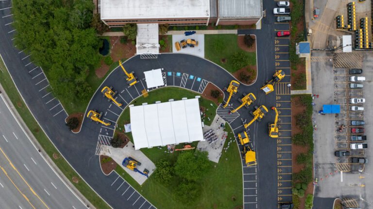 Aerial view of a construction yard with several yellow cranes surrounding a white tent on a curved asphalt driveway near a building and parking lot.