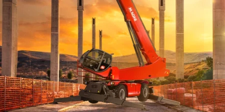 Red telescopic boom crane at a construction site with extended outriggers and orange safety fencing, set against a sunset sky and tall concrete pillars.