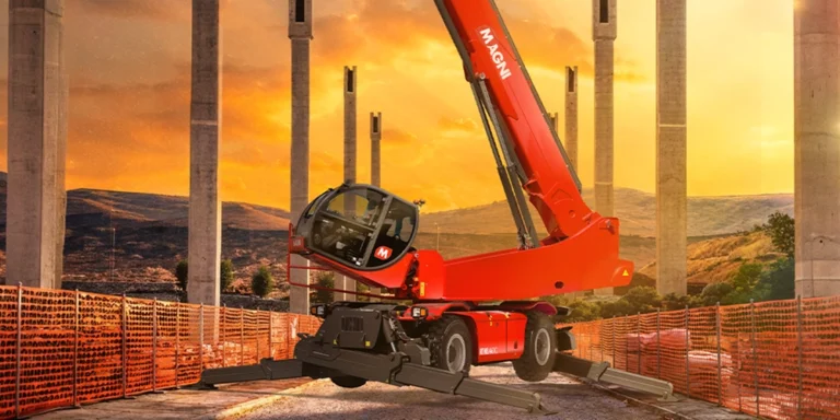 Red telescopic boom crane at a construction site with extended outriggers and orange safety fencing, set against a sunset sky and tall concrete pillars.