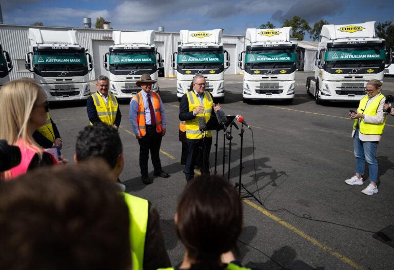Officials in high-visibility vests hold a press conference beside row of white Linfox trucks with 'Australian Made' banners in the background.