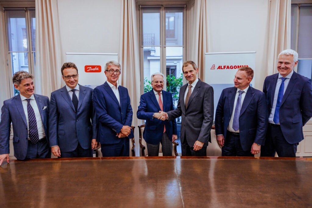 Danfoss Moves to Scale Up in Fluid Conveyance With Alfagomma Acquisition Seven businessmen in suits pose indoors; two center figures shake hands in front of company banners (Danfoss and ALFAGOMM).