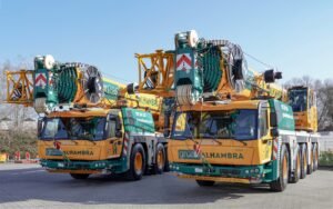 Gruas-Alhambra-adds-new-Grove-GMK5120L-cranes-at-SMOPYC-2026-01-scaledjpg - Machines Wiki Two large mobile crane trucks parked side by side, with reels, cables, and machinery mounted on their yellow and teal frames against a clear blue sky.