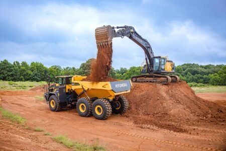 Hyundai-HX210L-loading-HA45-ADT_July-2026_mr-scaledjpg - Machines Wiki Excavator loads soil into a yellow Hyundai dump truck at a construction site on bare red earth.