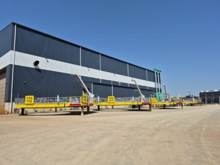 PALFINGER-PF-160-5m-davit-cranes-for-the-East-Anglia-TWO-offshore-wind-farmjpeg - Machines Wiki Industrial building with blue corrugated metal siding and a bright yellow elevated platform in front, under a clear sky.