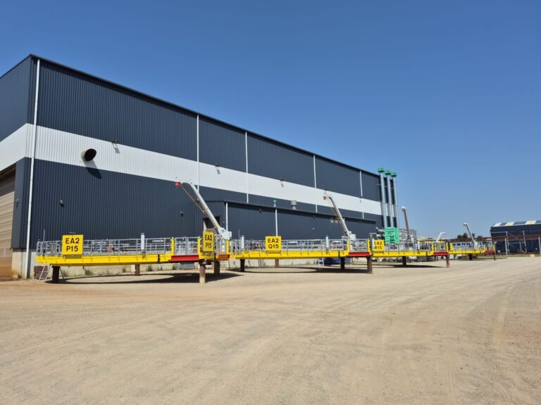 PALFINGER-PF-160-5m-davit-cranes-for-the-East-Anglia-TWO-offshore-wind-farmjpeg - Machines Wiki Industrial building with blue corrugated metal siding and a bright yellow elevated platform in front, under a clear sky.