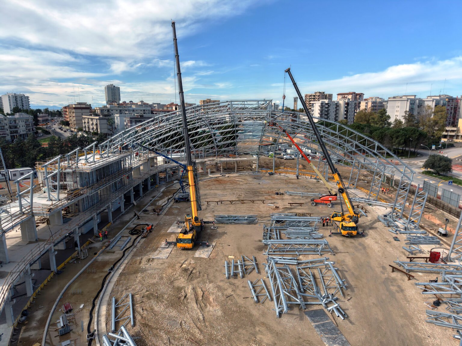 Two-Grove-cranes-power-construction-of-new-arena-for-the-Mediterranean-Games-2026_01-scaledjpg - Machines Wiki Construction site with steel lattice framework, cranes, and workers assembling a large building in a city background.