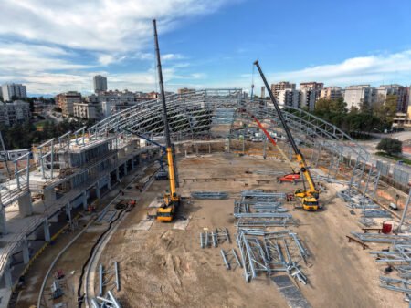 Two-Grove-cranes-power-construction-of-new-arena-for-the-Mediterranean-Games-2026_01-scaledjpg - Machines Wiki Construction site with steel lattice framework, cranes, and workers assembling a large building in a city background.