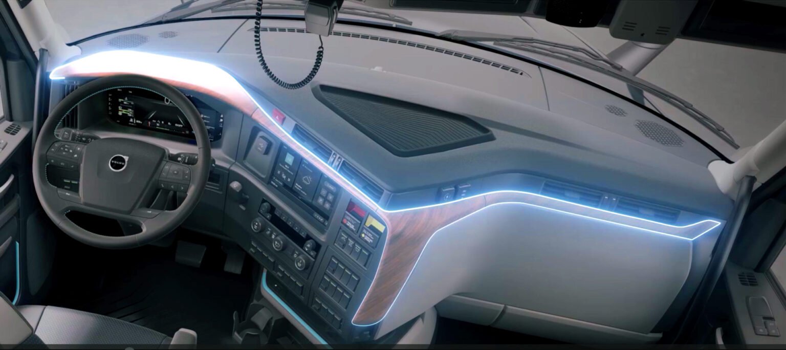 Volvo-ACT-Expo-2026-Sustainable-Materials-Video-1jpg - Machines Wiki Interior of a modern Volvo truck cabin with a digital instrument cluster, steering wheel, and wood-trim dash accented by blue ambient lights.