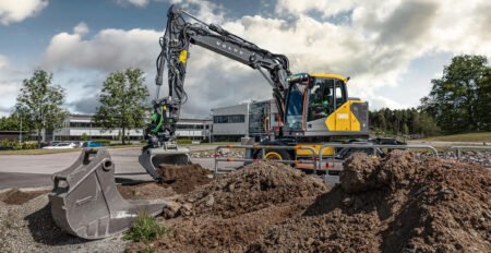 Large excavator with a long articulated arm digging at a construction site, dirt and rocks piled in the foreground and a modern office building in the background.