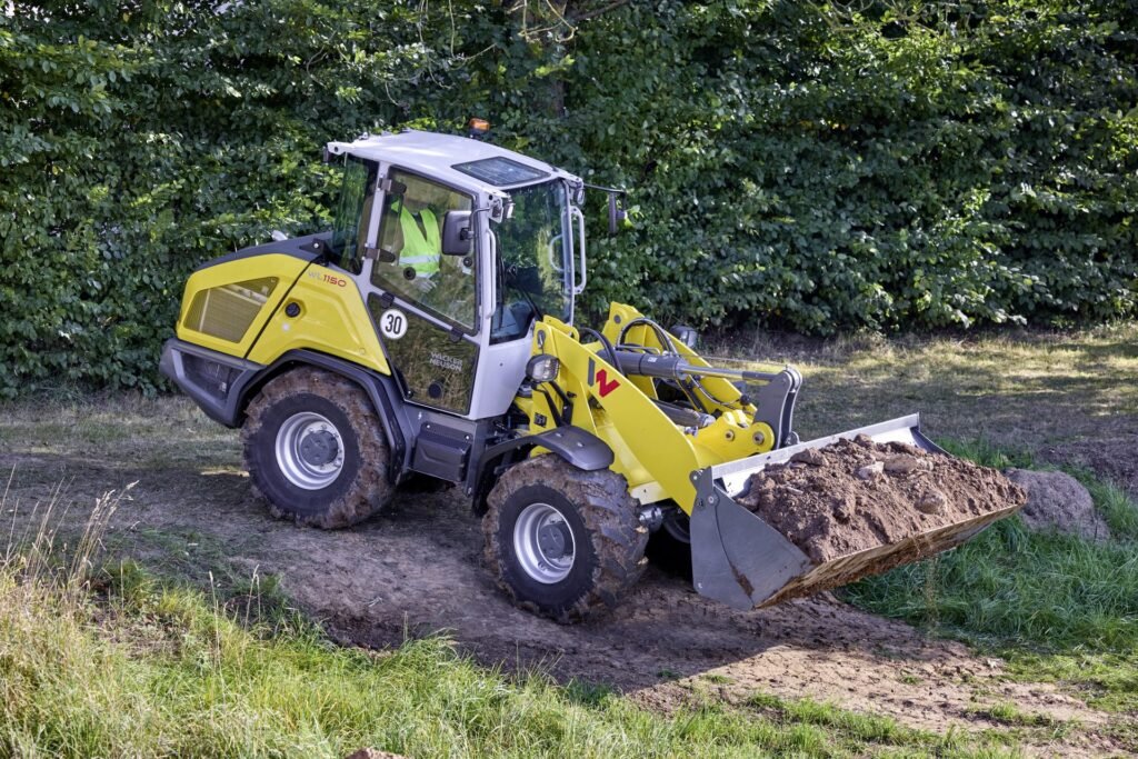 Yellow compact backhoe loader at a dirt site, with its front bucket filled with soil and green hedge in the background.