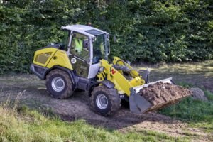 WN_PGK_image_WL1150_4_in_1_Schaufel_12A2439_action-copy-scaledjpg - Machines Wiki Yellow compact backhoe loader at a dirt site, with its front bucket filled with soil and green hedge in the background.