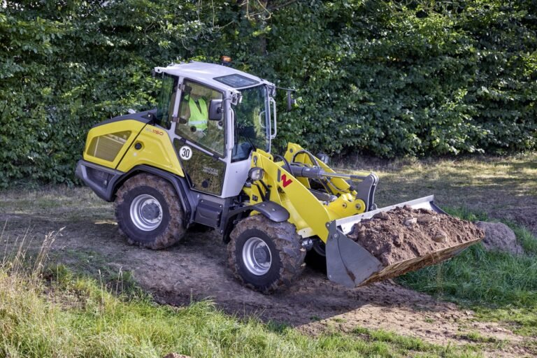 WN_PGK_image_WL1150_4_in_1_Schaufel_12A2439_action-copy-scaledjpg - Machines Wiki Yellow compact backhoe loader at a dirt site, with its front bucket filled with soil and green hedge in the background.