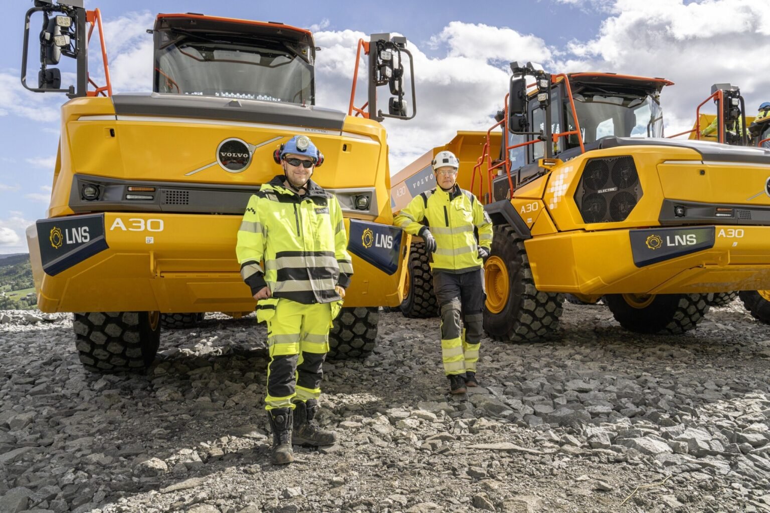 a30-electric-delivery-1-scaledjpg - Machines Wiki Two workers in high-visibility jackets stand in front of large yellow Volvo A30 dump trucks on rocky ground at a construction site.