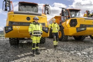 a30-electric-delivery-1-scaledjpg - Machines Wiki Two workers in high-visibility jackets stand in front of large yellow Volvo A30 dump trucks on rocky ground at a construction site.