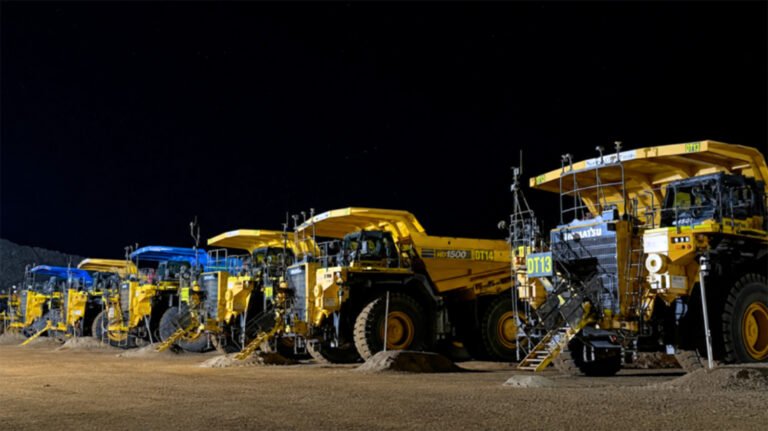 Row of large yellow mining dump trucks lined up at night on a dirt lot, with dark sky above.