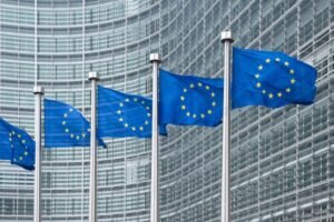 EU flags fluttering on flagpoles in front of a modern glass office building.